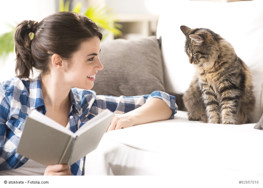 Eine Frau hält ein aufgeschlagenes Buch in der Hand und lacht ihre Katze an, die auf dem Sofa sitzt