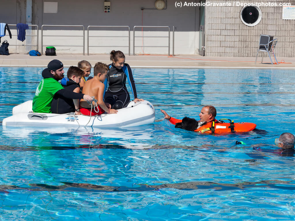 Schwimmbadtraining, Menschengruppe in Schlauchboot, Rettungsschwimmer mit Hund im Wasser.